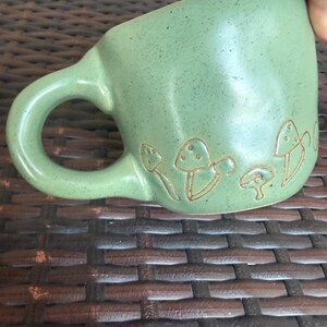 May include: A green ceramic mug with a rounded handle and a series of etched mushroom designs around the base. The mug has a speckled texture and a smooth, glossy finish. The mug is held by a hand.