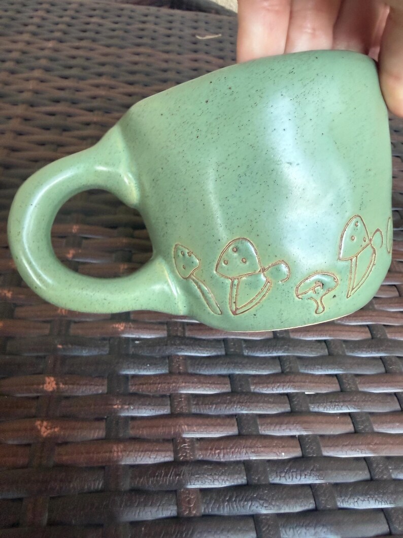 May include: A green ceramic mug with a rounded handle and a series of etched mushroom designs around the base. The mug has a speckled texture and a smooth, glossy finish. The mug is held by a hand.