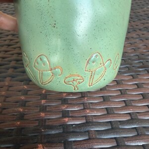 May include: A light green ceramic mug with a textured surface and a brown mushroom design around the base. The mug is held by a hand and is set against a brown woven background. The mug is approximately 4 inches tall.