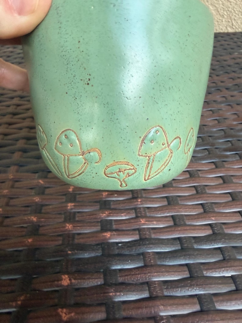 May include: A light green ceramic mug with a textured surface and a brown mushroom design around the base. The mug is held by a hand and is set against a brown woven background. The mug is approximately 4 inches tall.