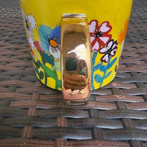 May include: A yellow ceramic mug with a white interior and a gold-colored handle. The mug is decorated with colorful floral designs in blue, red, and white. The mug is sitting on a brown woven surface.