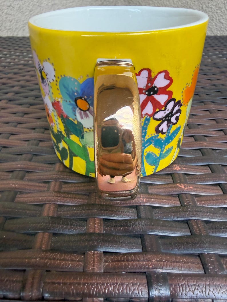 May include: A yellow ceramic mug with a white interior and a gold-colored handle. The mug is decorated with colorful floral designs in blue, red, and white. The mug is sitting on a brown woven surface.
