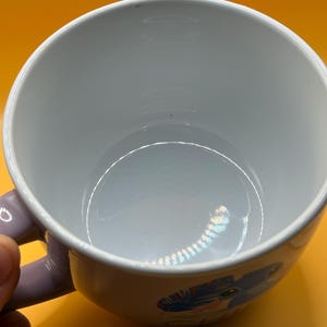 May include: White ceramic mug with a light purple handle. The mug features a colorful cartoon design on the side. The interior of the mug is white, and the background is yellow.