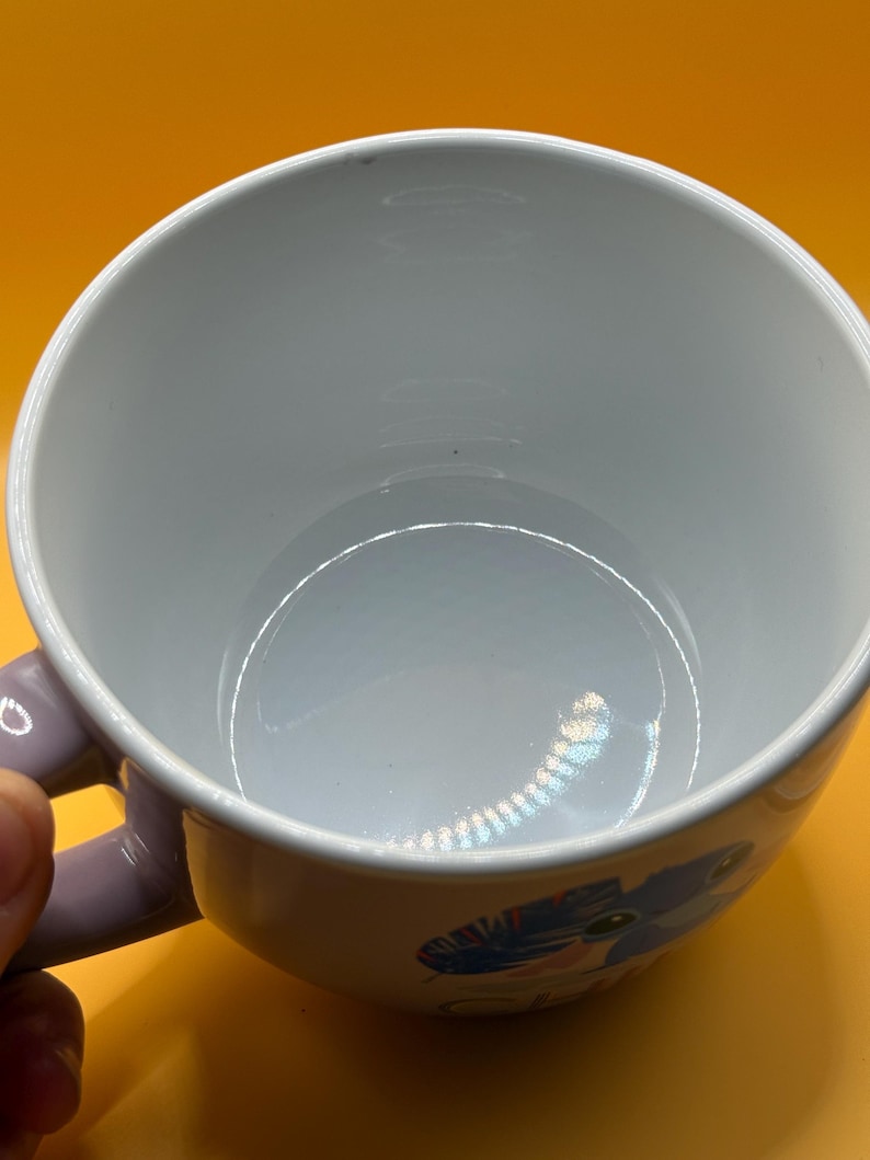 May include: White ceramic mug with a light purple handle. The mug features a colorful cartoon design on the side. The interior of the mug is white, and the background is yellow.