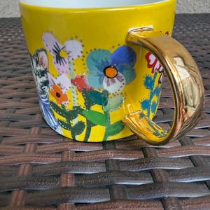May include: Yellow ceramic mug with a gold handle and a white interior. The mug is decorated with a floral design featuring blue, red, and orange flowers. A whimsical illustration of a person is also on the mug.