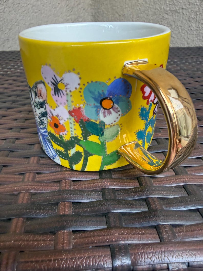 May include: Yellow ceramic mug with a gold handle and a white interior. The mug is decorated with a floral design featuring blue, red, and orange flowers. A whimsical illustration of a person is also on the mug.
