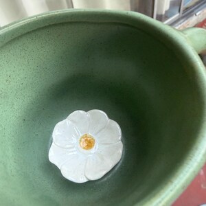 May include: A close-up shot of a green ceramic bowl with a white flower-shaped accent in the center. The flower has a yellow center. The bowl has a handle and a speckled texture.