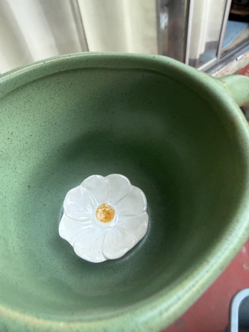 May include: A close-up shot of a green ceramic bowl with a white flower-shaped accent in the center. The flower has a yellow center. The bowl has a handle and a speckled texture.