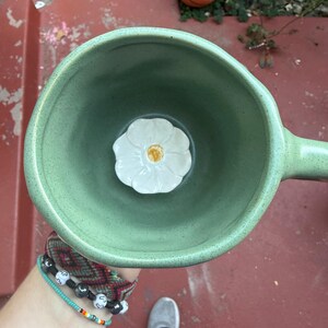 May include: A handmade, light green ceramic mug with a handle. Inside the mug is a white flower with a yellow center. The mug is held from a top-down perspective. A basketball is visible in the background.
