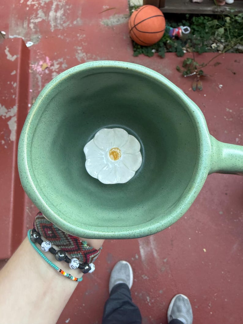 May include: A handmade, light green ceramic mug with a handle. Inside the mug is a white flower with a yellow center. The mug is held from a top-down perspective. A basketball is visible in the background.