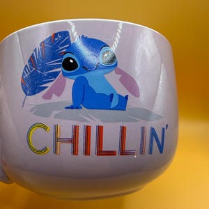 May include: A light purple mug featuring the character Stitch from Lilo & Stitch. Stitch is depicted in blue, with the word "CHILLIN'" in colorful letters. The mug has a handle and is set against a yellow background.