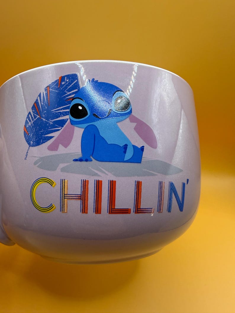 May include: A light purple mug featuring the character Stitch from Lilo & Stitch. Stitch is depicted in blue, with the word "CHILLIN'" in colorful letters. The mug has a handle and is set against a yellow background.