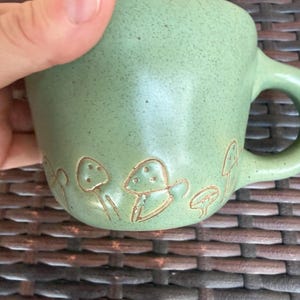 May include: A light green ceramic mug with a rounded handle. The mug features a series of embossed mushroom designs around the base. The mug has a speckled texture and a smooth, glossy finish.