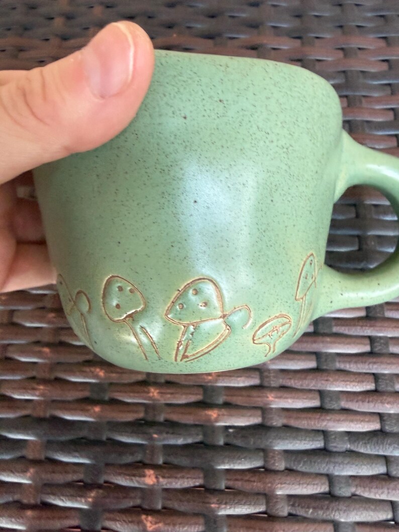 May include: A light green ceramic mug with a rounded handle. The mug features a series of embossed mushroom designs around the base. The mug has a speckled texture and a smooth, glossy finish.