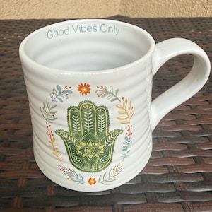 May include: White ceramic mug with a handle, featuring a green Hamsa hand design surrounded by colorful floral accents. The mug has the text "Good Vibes Only" in light blue near the rim. The mug has a ribbed texture.