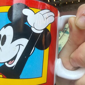May include: A white ceramic mug featuring a graphic of Mickey Mouse with black ears, a white face, and a red shirt. The character is set against a blue background framed by a yellow border, all enclosed within a vibrant red field on the mug surface.