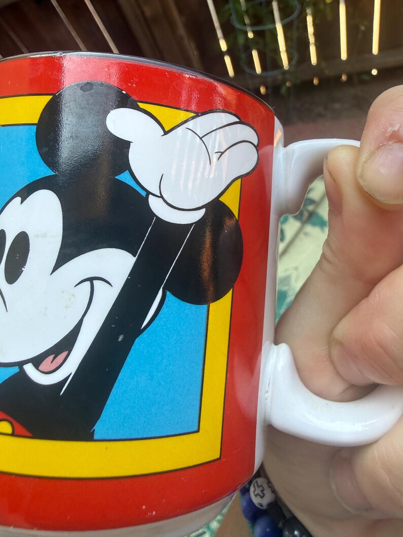 May include: A white ceramic mug featuring a graphic of Mickey Mouse with black ears, a white face, and a red shirt. The character is set against a blue background framed by a yellow border, all enclosed within a vibrant red field on the mug surface.