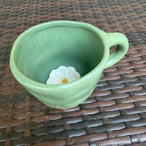 May include: A handmade, green ceramic mug with a rounded shape and a handle. Inside the mug, a white flower with a yellow center is placed. The mug is sitting on a brown woven surface.