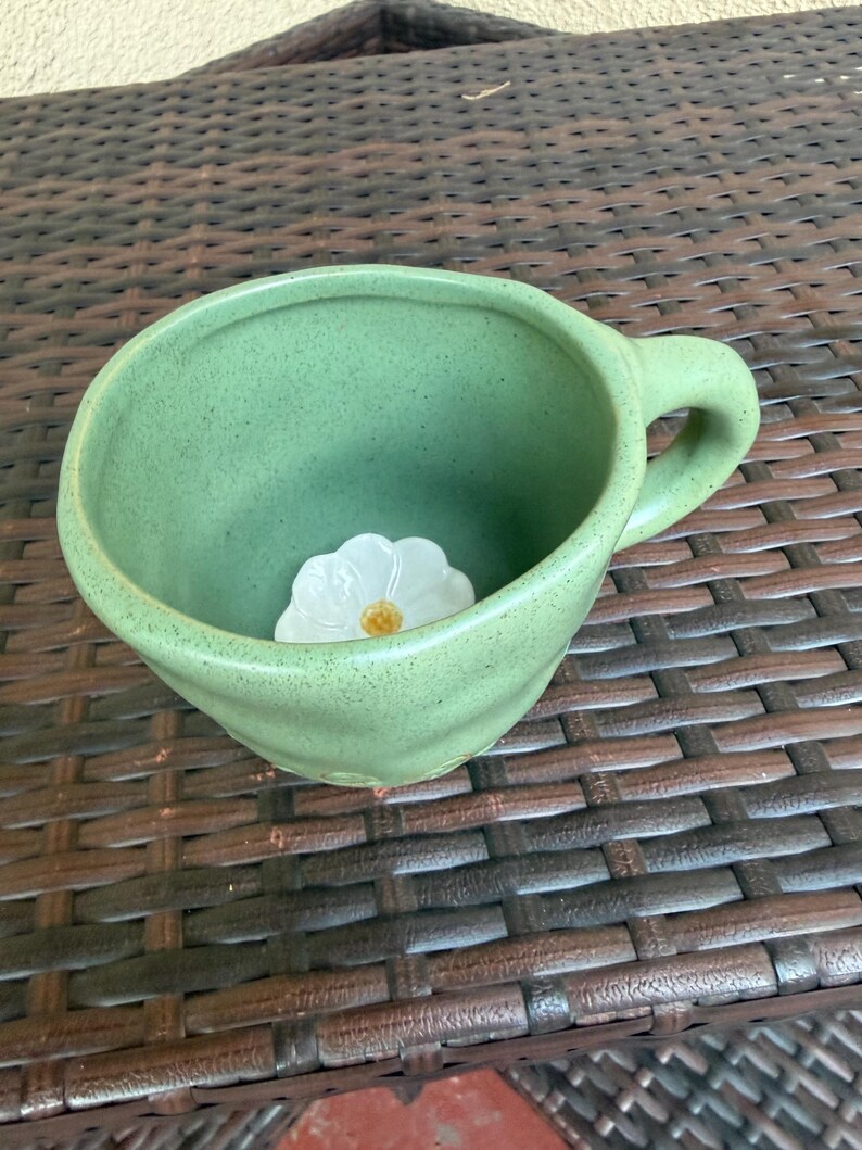 May include: A handmade, green ceramic mug with a rounded shape and a handle. Inside the mug, a white flower with a yellow center is placed. The mug is sitting on a brown woven surface.