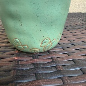 May include: A handmade, light green ceramic mug with a textured surface. The mug features embossed mushroom designs near the base. The mug is set against a dark brown woven background, creating a rustic aesthetic.