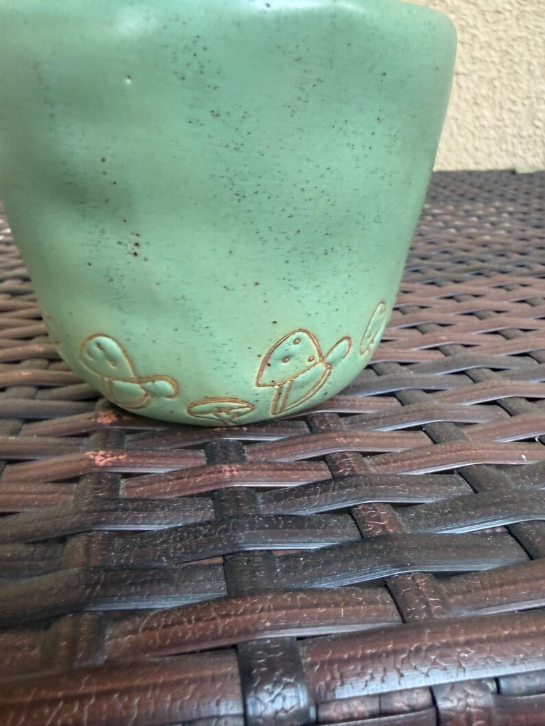 May include: A handmade, light green ceramic mug with a textured surface. The mug features embossed mushroom designs near the base. The mug is set against a dark brown woven background, creating a rustic aesthetic.