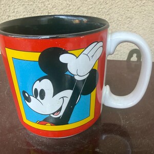 May include: A ceramic mug with a red exterior and a black interior. The front features a graphic of Mickey Mouse waving, set within a blue square bordered by a yellow frame. The mug has a white handle and a smooth, glossy finish throughout.