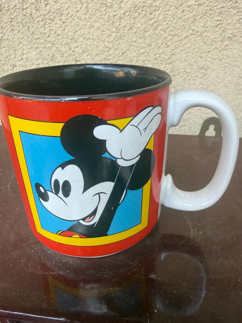May include: A ceramic mug with a red exterior and a black interior. The front features a graphic of Mickey Mouse waving, set within a blue square bordered by a yellow frame. The mug has a white handle and a smooth, glossy finish throughout.