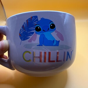 May include: A lavender ceramic mug featuring a blue cartoon character with large black eyes and pink ears. The word "CHILLIN" is printed in colorful letters below the character. The mug has a handle on the left side.