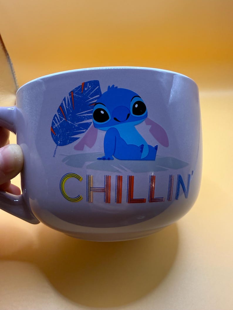 May include: A lavender ceramic mug featuring a blue cartoon character with large black eyes and pink ears. The word "CHILLIN" is printed in colorful letters below the character. The mug has a handle on the left side.