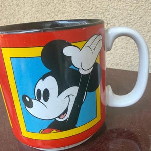 May include: A ceramic mug with a red exterior and a black interior. The front features a graphic of Mickey Mouse waving, set within a blue square bordered by a yellow frame. The mug has a white handle and a white base, showcasing a classic design.