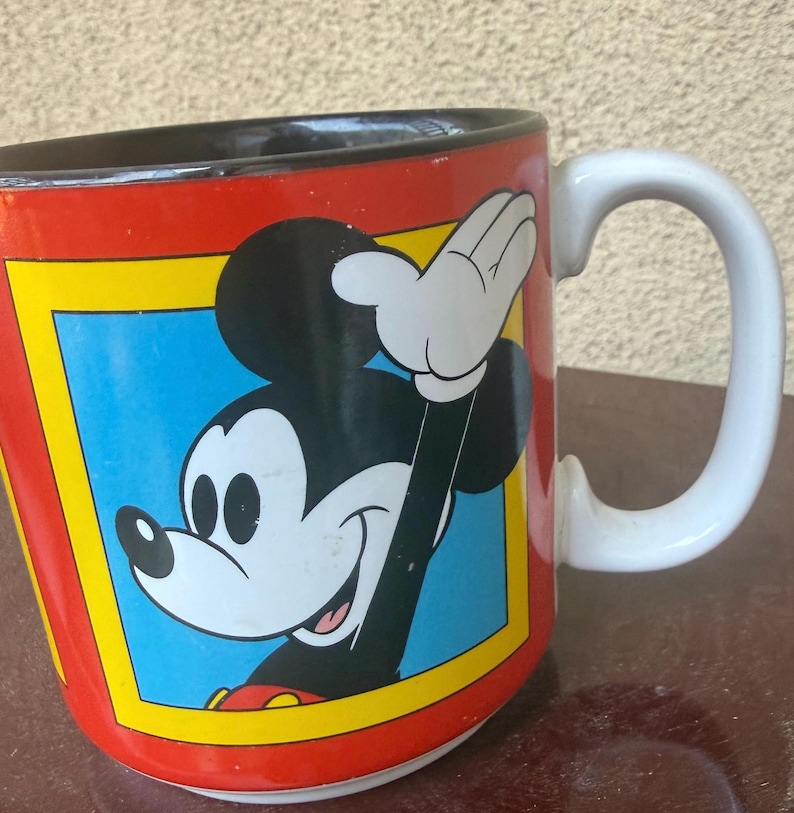 May include: A ceramic mug with a red exterior and a black interior. The front features a graphic of Mickey Mouse waving, set within a blue square bordered by a yellow frame. The mug has a white handle and a white base, showcasing a classic design.
