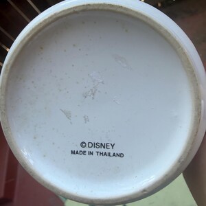 May include: The circular white base of a ceramic object features the black printed text "&copy;DISNEY" and "MADE IN THAILAND" centered on the bottom. The underside has a slightly raised rim and shows minor signs of wear with light brown discoloration.