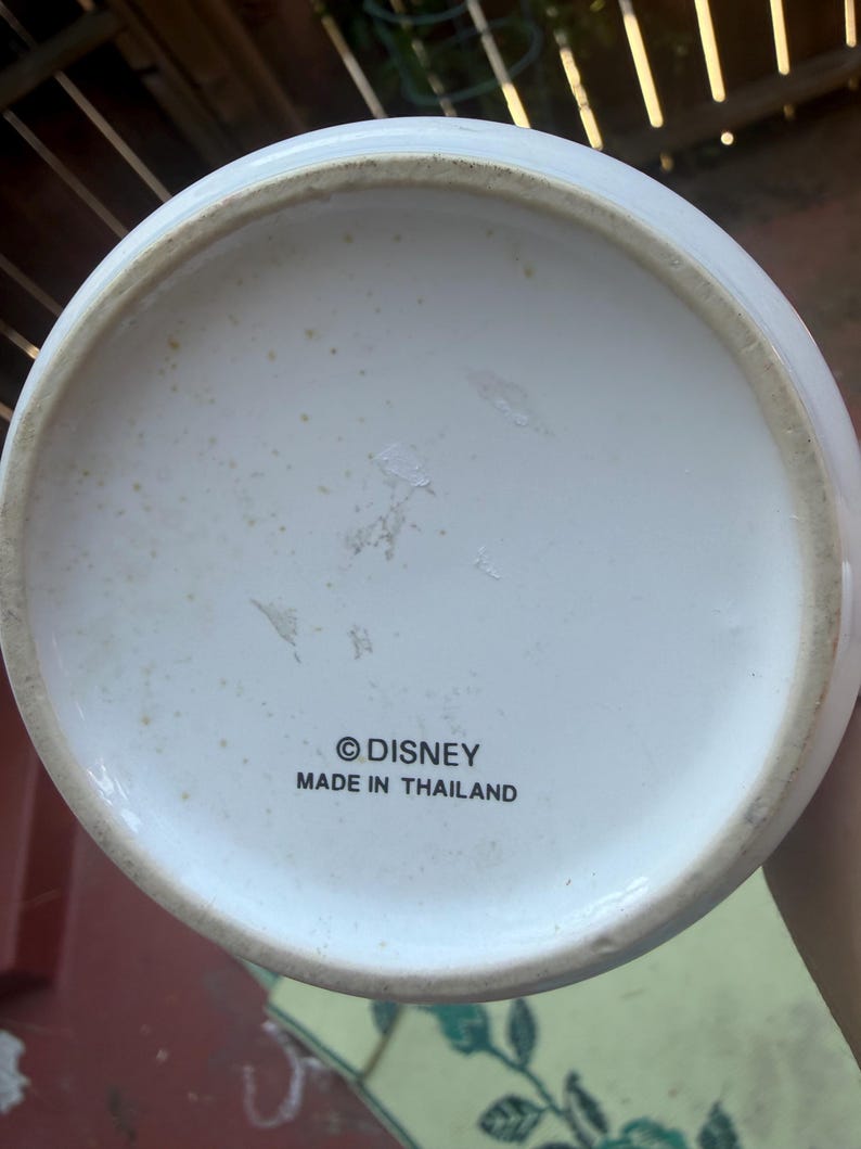 May include: The circular white base of a ceramic object features the black printed text "&copy;DISNEY" and "MADE IN THAILAND" centered on the bottom. The underside has a slightly raised rim and shows minor signs of wear with light brown discoloration.