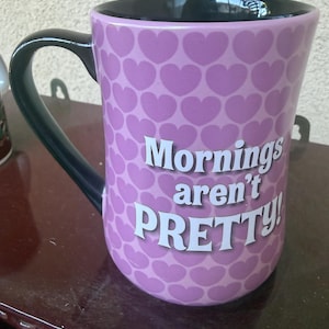 Puede incluir: Una taza de cer&aacute;mica color lavanda con un patr&oacute;n repetitivo de corazones de color p&uacute;rpura oscuro en el exterior. El asa y el interior de la taza son negros. El texto "Mornings aren't PRETTY!" est&aacute; impreso en blanco en la parte frontal.