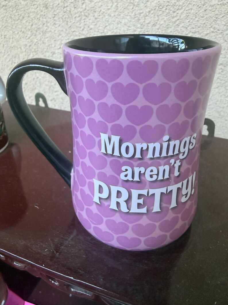 Puede incluir: Una taza de cer&aacute;mica color lavanda con un patr&oacute;n repetitivo de corazones de color p&uacute;rpura oscuro en el exterior. El asa y el interior de la taza son negros. El texto "Mornings aren't PRETTY!" est&aacute; impreso en blanco en la parte frontal.