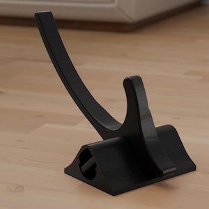 May include: A black, curved display stand, likely for a sword or similar item. The stand has a wide, rectangular base with a curved support that rises and curves back down. The stand is made of a solid, matte black material.