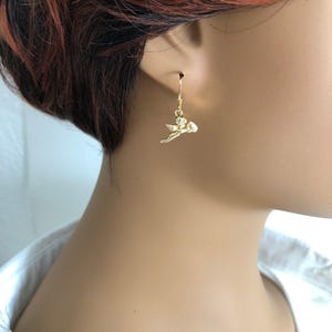 May include: Gold-colored dangle earrings featuring a small cherub design. The earrings have a hook closure and a detailed cherub pendant with outstretched wings. The cherub is holding a small heart. The earrings are shown on a person's ear.