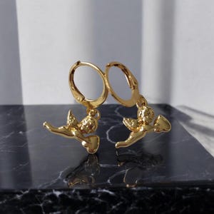 May include: A pair of gold-colored hoop earrings. Each earring features a small, detailed cherub pendant holding a bow and arrow. The earrings are displayed on a dark, marbled surface, with a white background.