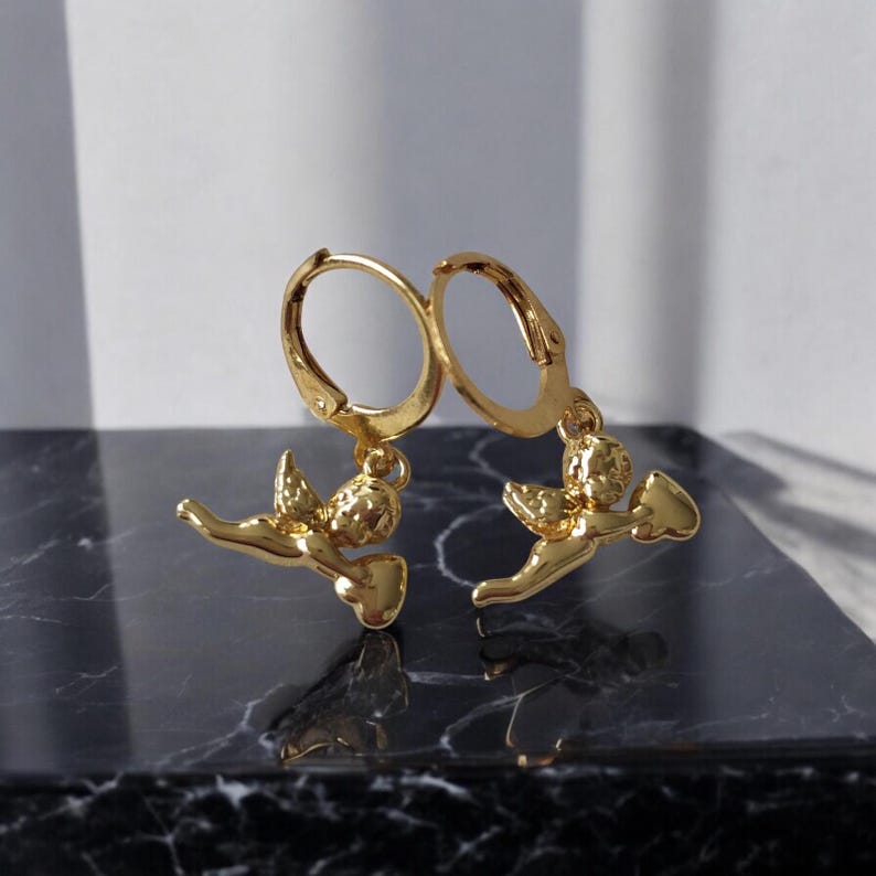 May include: A pair of gold-colored hoop earrings. Each earring features a small, detailed cherub pendant holding a bow and arrow. The earrings are displayed on a dark, marbled surface, with a white background.