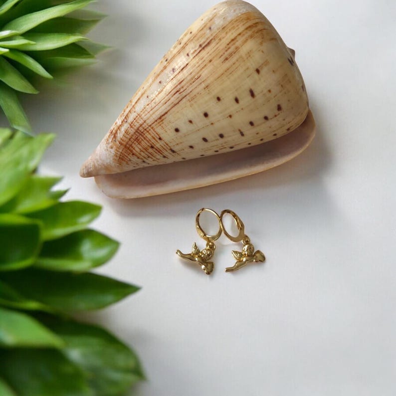 May include: A pair of gold-colored hoop earrings with a bird-like charm, resting on a white surface. A large, tan and brown seashell is positioned behind the earrings. Green leafy plants are visible in the background.