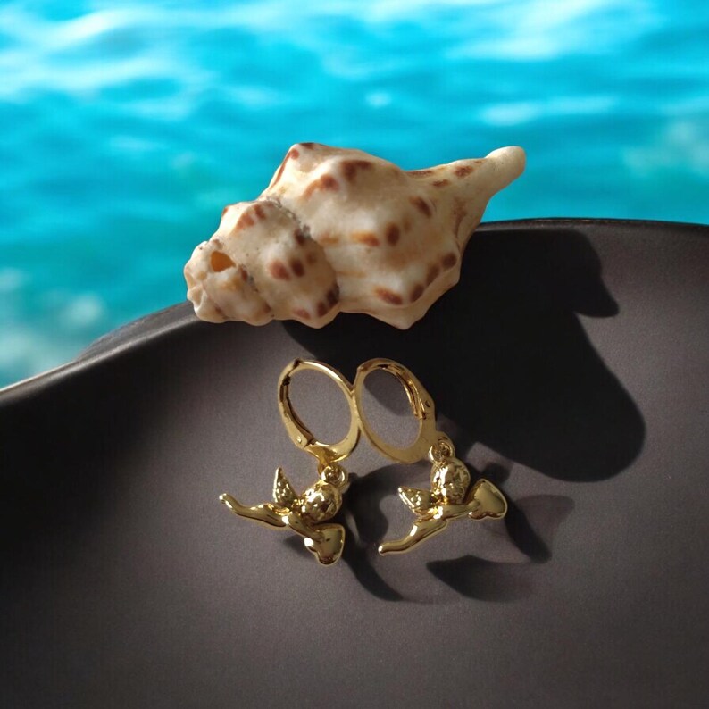 May include: A pair of gold-colored hoop earrings with a bird perched on a branch design. A seashell rests on a dark surface, with a blurred blue background. The earrings are detailed and shiny.