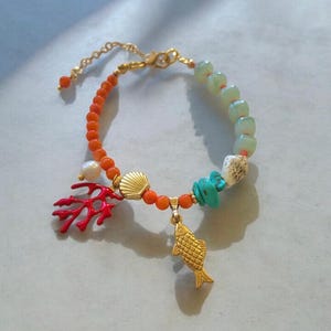 May include: A beaded bracelet featuring a mix of orange, pale green, and turquoise beads, accented with a white pearl, a gold-toned shell charm, a red coral-shaped charm, and a gold-toned fish charm, finished with a gold-toned metal chain clasp.
