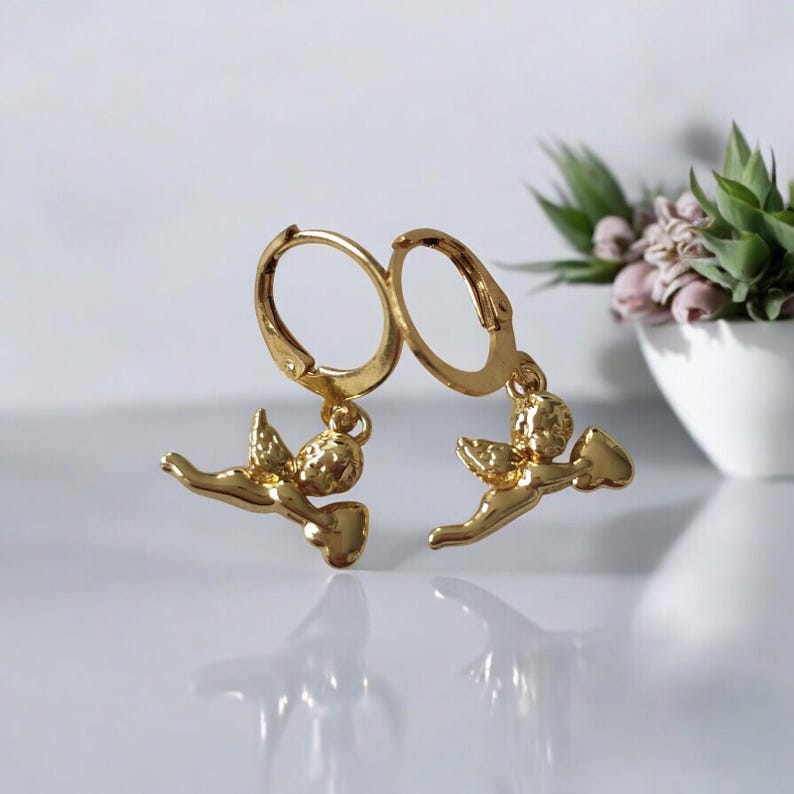 May include: A pair of gold-colored hoop earrings. Each earring features a small, detailed cherub pendant. The cherubs are depicted with wings and are holding a small object. The earrings are displayed on a white surface.