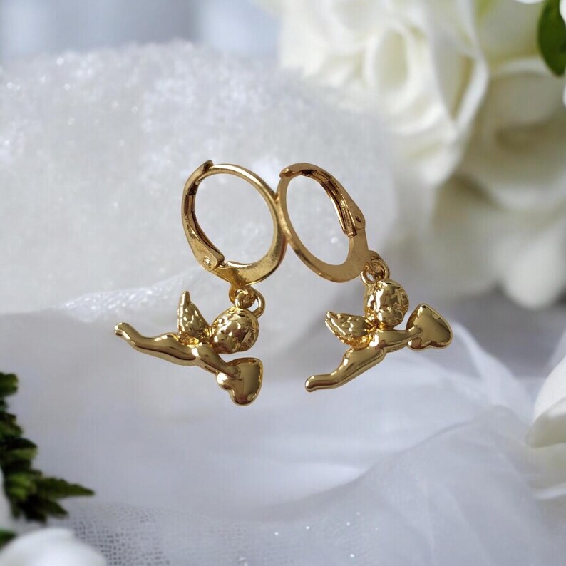 May include: A pair of gold-colored hoop earrings. Each earring features a small, detailed cherub pendant. The cherubs are in a flying pose, holding a heart-shaped object. The earrings are displayed on a white, textured surface.