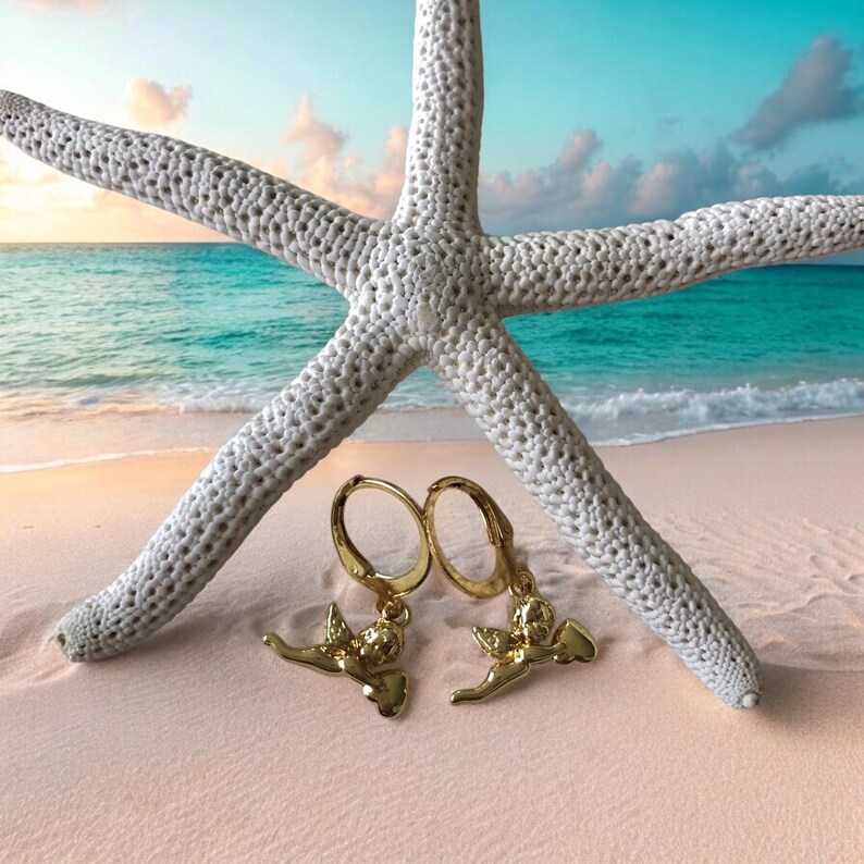 May include: Gold-colored hoop earrings with a cherub design, resting on a sandy beach with a white starfish and ocean backdrop. The earrings are detailed with a small cherub figure. The beach has pink sand and the ocean is turquoise.