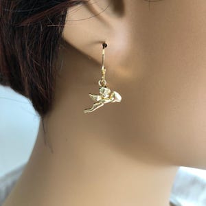 May include: Gold-colored angel earrings. Each earring features a small, detailed angel charm suspended from a lever-back closure. The angel is depicted in a flying pose, with wings and a small heart-shaped object.