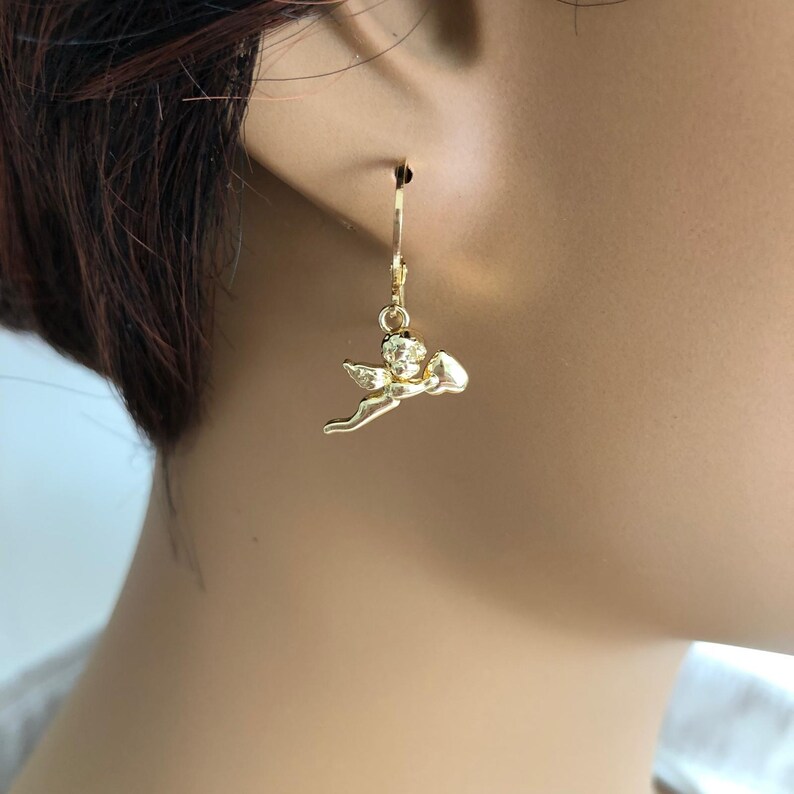 May include: Gold-colored angel earrings. Each earring features a small, detailed angel charm suspended from a lever-back closure. The angel is depicted in a flying pose, with wings and a small heart-shaped object.