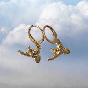 May include: A pair of gold-colored hoop earrings. Each earring features a small, detailed angel charm with outstretched wings and a flowing garment. The earrings are set against a backdrop of a cloudy sky.