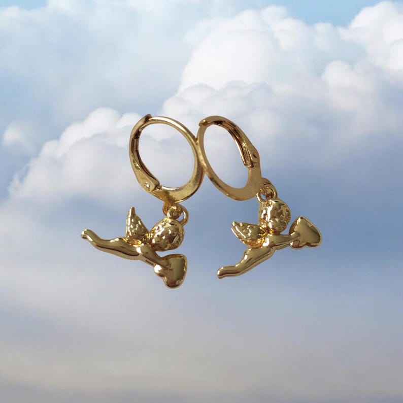 May include: A pair of gold-colored hoop earrings. Each earring features a small, detailed angel charm with outstretched wings and a flowing garment. The earrings are set against a backdrop of a cloudy sky.