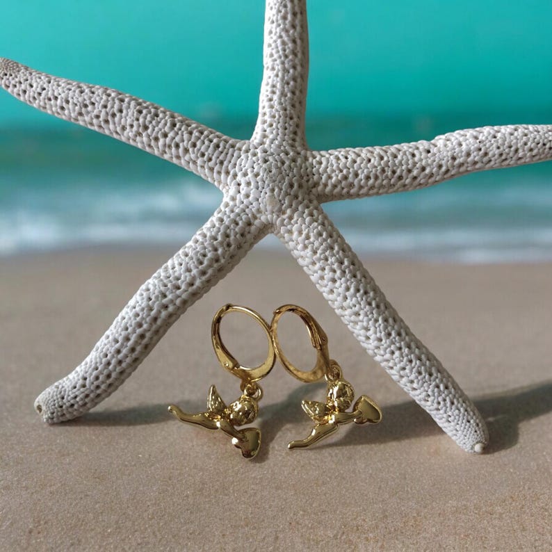 May include: A pair of gold-colored earrings with a cherub design, resting on a sandy surface. A large white starfish is in the background, with a turquoise ocean and beach in the distance. The earrings have a lever back closure.